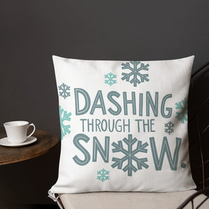 A pillow on a chair with a coffee mug on a table next to it. The lettering says "Dashing through the snow" in light and dark blue with snowflakes surrounding the words. The words and snowflakes create a pattern on the pillow. 
