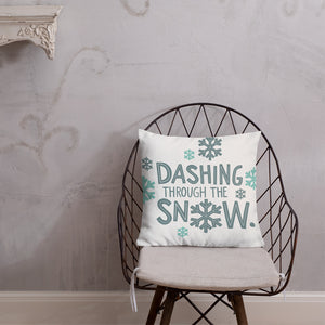 The pillow is leaning on a metal chair with a cushion. The white pillow has a repeat pattern with the phrase "dashing through the snow" in light and dark blue and snowflakes around the words. 