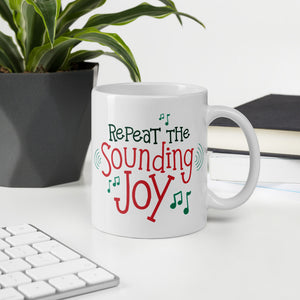 A mug featured on a desk with a plant and a keyboard. The illustrated design says "Repeat the Sounding Joy" with music notes around it. 