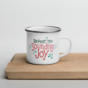A white enamel mug sitting on top of a wood cutting board on a white background. The mug is white with the very top silver enamel. The design features the words "Repeat the Sounding Joy" in red and green with musical notes around the letters. 