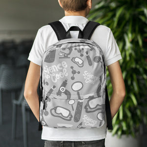 A boy with his back to the camera and dining chairs and a plant in the distance. The backpack is a light gray with a pattern of illustrations in darker gray and white. The pattern of illustrations features test tubes, microscopes, magnifying glasses, protective science goggles, atom models and the words "Science is cool."