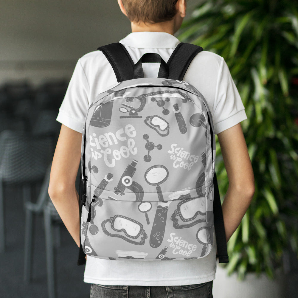 A boy with his back to the camera and dining chairs and a plant in the distance. The backpack is a light gray with a pattern of illustrations in darker gray and white. The pattern of illustrations features test tubes, microscopes, magnifying glasses, protective science goggles, atom models and the words 