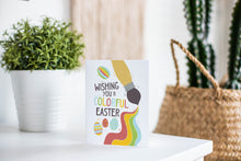 Load image into Gallery viewer, A greeting card is featured on a white tabletop with a white planter in the background with a green plant. There’s a woven basket in the background with a cactus inside. The card features an illustrated paint brush and Easter eggs with the words “Wishing you a colorful Easter.”