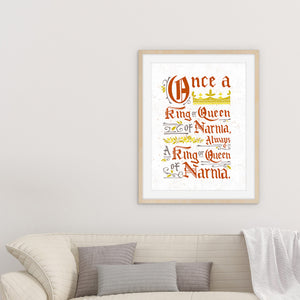 A light wood frame above a sofa. Artwork in a white frame with the with a white matte. The frame is leaning on a white counter. The artwork features hand drawn lettering of the Narnia quote "Once a king or queen of Narnia, always a king or queen of Narnia."