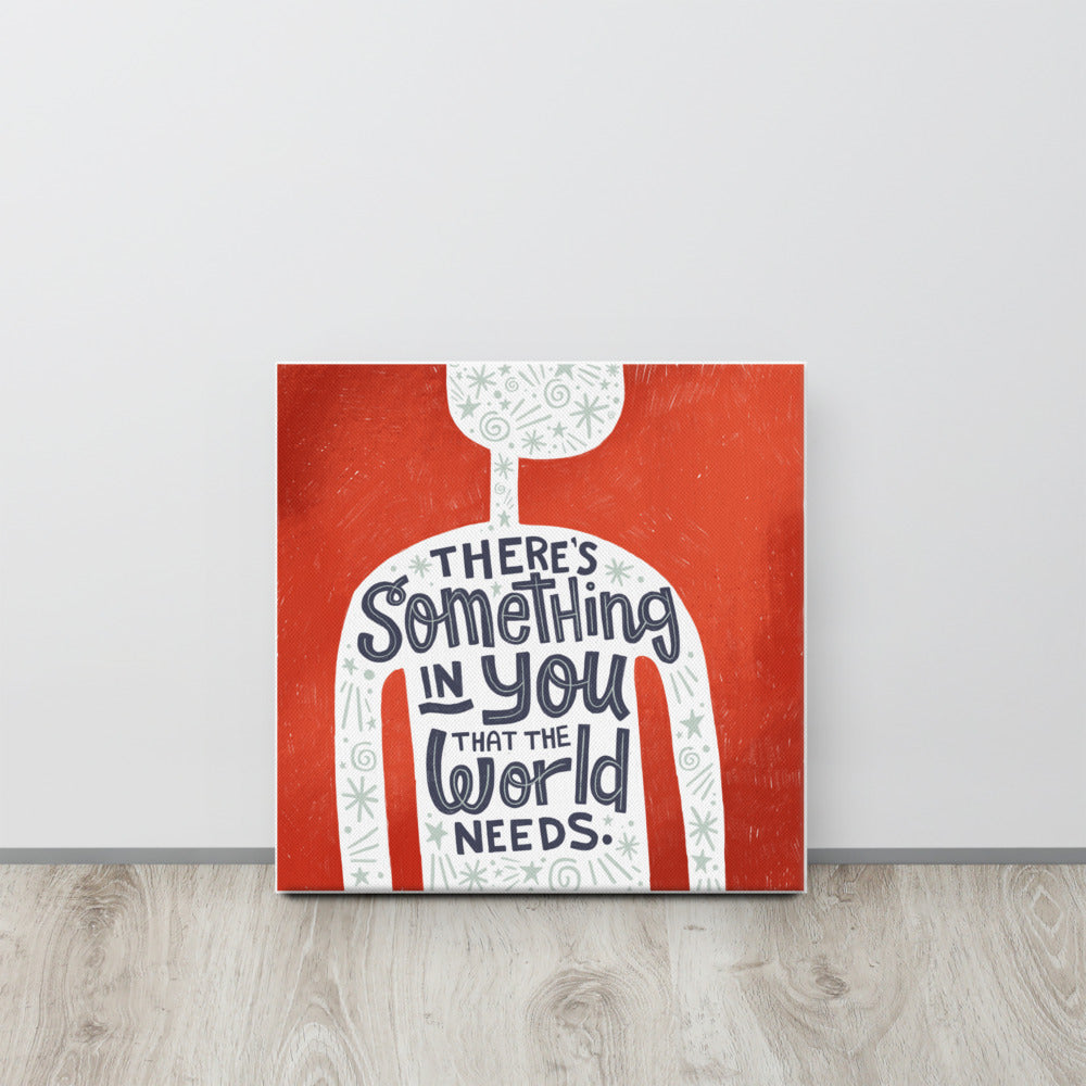 A red wall art canvas leans against a pale grey wall. The canvas design features the outline of a person in white, filled with light grey doodles. The words ‘There’s something in you the world needs' are lettered in black across the person’s chest.