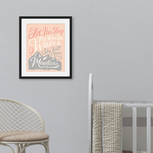 A pink print hanging on a pale grey wall, next to a white cot. The print reads 'Let her sleep for when she wakes she will move mountains' in a pink, white, and light grey lettering design, with a grey mountain illustration at the bottom.