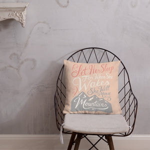 A pink cushion sits on a metal chair against a pale plaster wall. The cushion reads 'Let her sleep for when she wakes she will move mountains' in a pink, white, and light grey lettering design, with a grey mountain illustration at the bottom.