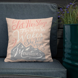A pink cushion sits on a dark sofa. The cushion reads 'Let her sleep for when she wakes she will move mountains' in a pink, white, and light grey lettering design, with a grey mountain illustration at the bottom.