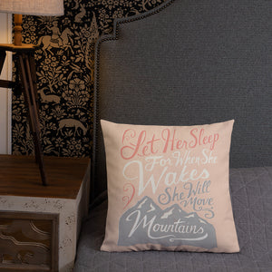 A pink cushion leands against a grey headboard next to a small lamp. The cushion reads 'Let her sleep for when she wakes she will move mountains' in a pink, white, and light grey lettering design, with a grey mountain illustration at the bottom.