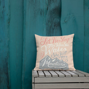 A pink cushion leans against a painted teal fence. The cushion reads 'Let her sleep for when she wakes she will move mountains' in a pink, white, and light grey lettering design, with a grey mountain illustration at the bottom.