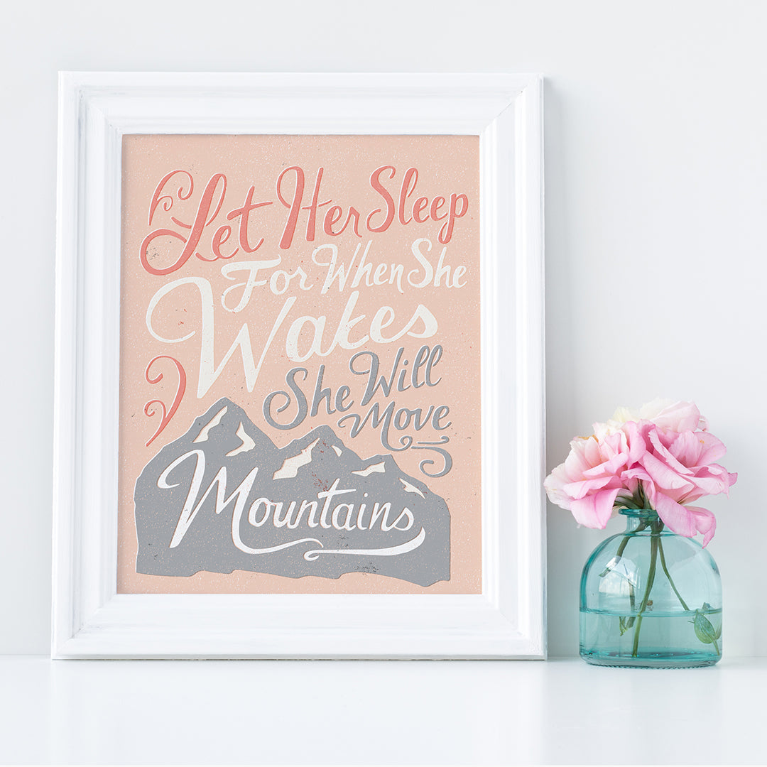 A pink print in a white frame sits beside a vase of pink flowers. The print reads 'Let her sleep for when she wakes she will move mountains' in a pink, white, and light grey lettering design, with a grey mountain illustration at the bottom.