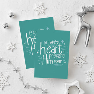 Two Christmas cards laying on a white background with white and silver Christmas decorations on the table. The card background color is teal with white lettering reading "Let every heart prepare him room" with stars and lines around the words.