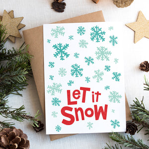 A Christmas card on top of a brown craft paper gift. The card has a white background with the words "let it snow" at the bottom in red. Illustrated snowflakes are on the rest of the card in different shades of blue.