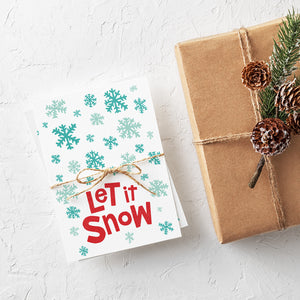 A stack of Christmas cards with brown string wrapped around them. A brown craft paper gift is off to the side. The card has a white background with the words "let it snow" at the bottom in red. Illustrated snowflakes are on the rest of the card in different shades of blue.