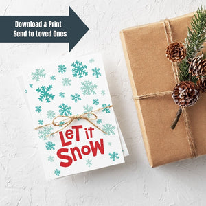 A stack of Christmas cards with brown string wrapped around them. A brown craft paper gift is off to the side. The card has a white background with the words "let it snow" at the bottom in red. Illustrated snowflakes are on the rest of the card in different shades of blue. The words "download & print, send to loved ones" are on the top lefthand part of the image. 