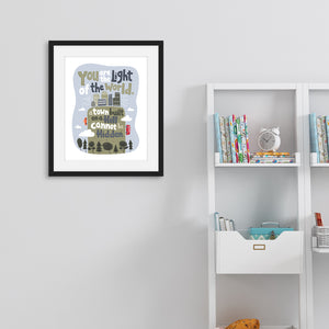 A black framed picture on a the wall with kids shelving on the side. The artwork is on a white background with lettering reading "You are the light of the world, a town built on a hill cannot be hidden." The words are a light gray background with an illustrated city.