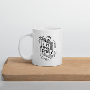 A white mug sitting on a light wood cutting board. The design features the words "Live Your Story" with the words inside an illustrated book. 