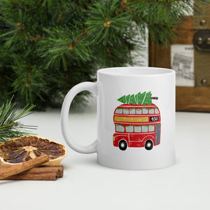 A white mug with a pine tree in the background. The mug feaures an illustrated London red double decker bug with a Christmas theme. The bus has a Christmas tree laying on the top and green garland in the windows. 