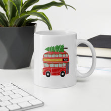 Load image into Gallery viewer, A mug featured on a desk with a plant and a keyboard. The white mug features hand drawn illustrated London bus. The red double decker bus has a Christmas tree laying on the top and Christmas garland in the bus windows. 