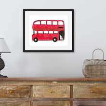 Load image into Gallery viewer, A black frame with an illustration of a red double decker bus. The frame is above a dresser with a basket and lamp on the dresser. 