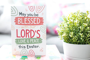 A photo of a card featured on a tabletop next to a white planter filled with a green plant. ​​The card features the words “May You be Blessed with our Lord's Love & Peace this Easter.”