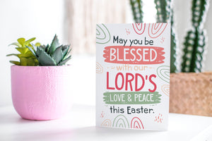 A greeting card featured standing up on a white tabletop with a pink plant pot in the background and some succulents in the pot. There’s a woven basket in the background with a cactus inside. The card features the words “May You be Blessed with our Lord's Love & Peace this Easter.”