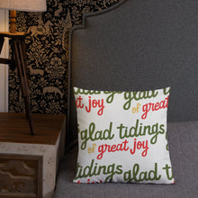 Load image into Gallery viewer, A pillow leaning on a grey headboard with a table and lamp off to the side. The white pillow is white with the words &quot;glad tidings of great joy&quot; in green, red and yellow. The words create a pattern on the pillow. 