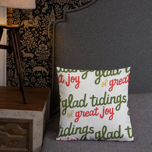A pillow leaning on a grey headboard with a table and lamp off to the side. The white pillow is white with the words "glad tidings of great joy" in green, red and yellow. The words create a pattern on the pillow. 
