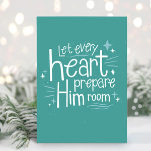 A Christmas card standing up with with pine leaves in the background with a touch of snow. The card background color is teal with white lettering reading "Let every heart prepare him room" with stars and lines around the words. 