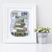 Load image into Gallery viewer, Artwork in a white frame with the with a white matte. The frame is leaning on a shelf with a metal plant pot next to it with white flowers. The artwork is on a white background with lettering reading &quot;You are the light of the world, a town built on a hill cannot be hidden.&quot; The words are a light gray background with an illustrated city.