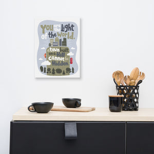 A canvas is hanging on a wall above a kitchen counter with coffee mugs and kitchen utensils. The artwork is on a white background with lettering reading "You are the light of the world, a town built on a hill cannot be hidden." The words are a light gray background with an illustrated city.