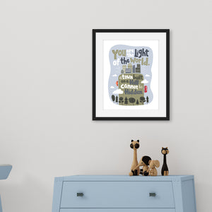 A black frame of illustrated artwork above a light blue children's dresser with kids toys on the top. The artwork is on a white background with lettering reading "You are the light of the world, a town built on a hill cannot be hidden." The words are a light gray background with an illustrated city.