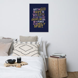 A canvas hanging above a bed. The canvas is purple and features hand drawn text in white, pink and yellow reading "May the God of hope fill you with all joy and peace as you trust him, so that you may overflow with hope by the power of the holy spirit." 