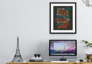 Artwork in a black frame with the with a white matte shown hanging on a wall above a desk. The artwork is on a black background with colorful letters reading May the God of hope fill you with all joy and peace as you trust him, so that you may overflow with hope by the power of the holy spirit."