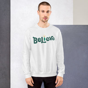 A man wearing a white sweatshirt featuring hand drawn lettering with the word Believe in green. The "I" of the word Believe is an illustrated Christmas tree. 