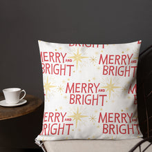 Load image into Gallery viewer, A pillow on a chair with a coffee mug on a table next to it. The white pillow features the words merry and bright in red and yellow Christmas stars in a pattern. 