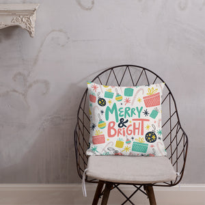 The pillow is leaning on a metal chair with a cushion. The white pillow features a Christmas pattern with gifts, ornaments, stars and candy canes. The words Merry & Bright are in the center of the pillow. The colors of the pattern and words are light blue, yellow, pink and black. 