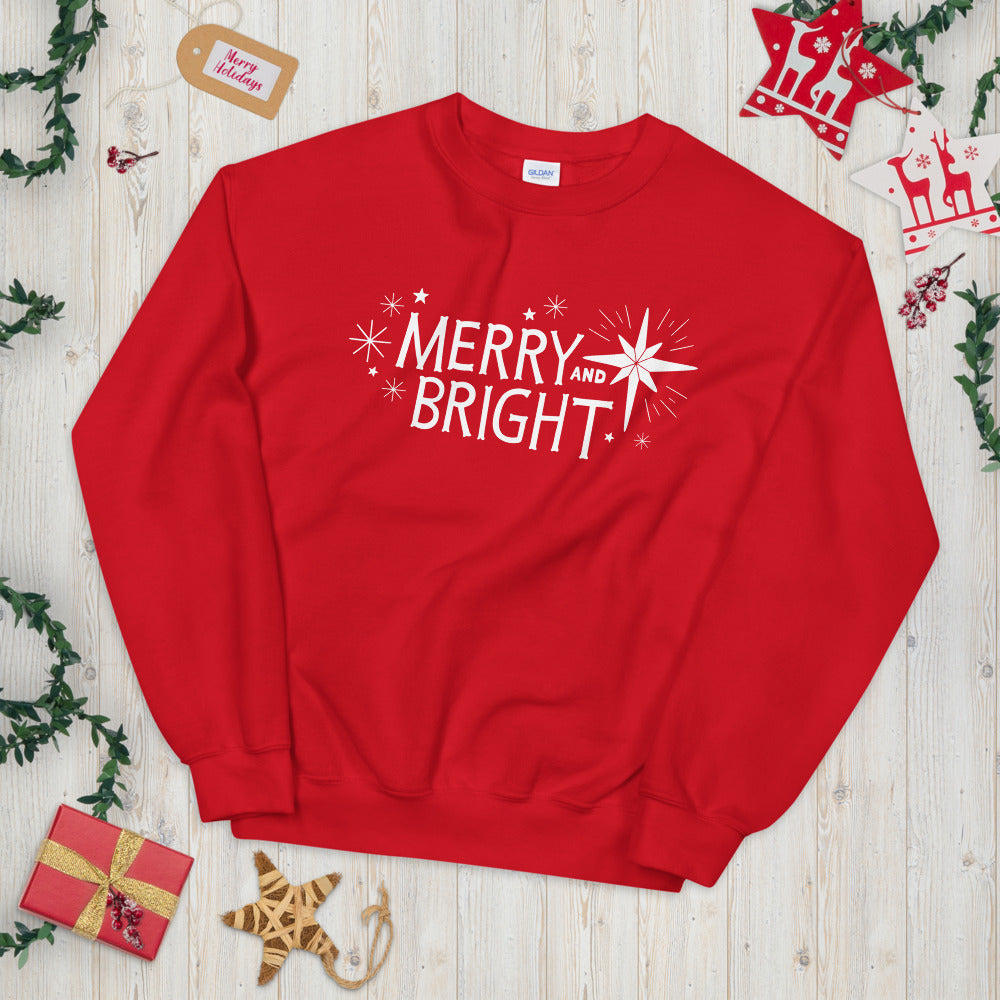 A red sweatshirt laying on a table with Christmas objects around it. The sweatshirt features the words Merry and Bright with illustrated Christmas stars around it. The words and illustrations are in white on the red fabric. 