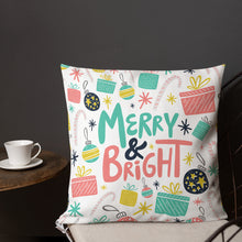 Load image into Gallery viewer, A pillow on a chair with a coffee mug on a table next to it. The white pillow features Christmas illustrations of gifts, ornaments, stars and candy canes. The words Merry &amp; Bright are in the middle of the pillow and the words and pattern are in the colors light blue, pink, yellow and black. 
