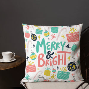 A pillow on a chair with a coffee mug on a table next to it. The white pillow features Christmas illustrations of gifts, ornaments, stars and candy canes. The words Merry & Bright are in the middle of the pillow and the words and pattern are in the colors light blue, pink, yellow and black. 