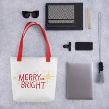 Load image into Gallery viewer, A tote bag lying on a surface with a laptop and office items next to it. The patterned tote bag has the words Merry and Bright in red and yellow illustrated stars. The handles are red. 
