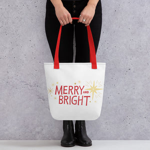 Someone holding a tote bag with red handles and a white fabric bag. The artwork features an illustrated Christmas pattern with yellow stars and the words Merry and Bright in red. 