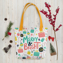 Load image into Gallery viewer, A white tote bag with yellow handles laying on a table with Christmas items around it. The tote bag features illustrated presents, ornaments, stars and candy canes. The words Merry &amp; Bright are in the center of the pattern. The colors of the illustrated patterns are pink, light blue, yellow and black. 