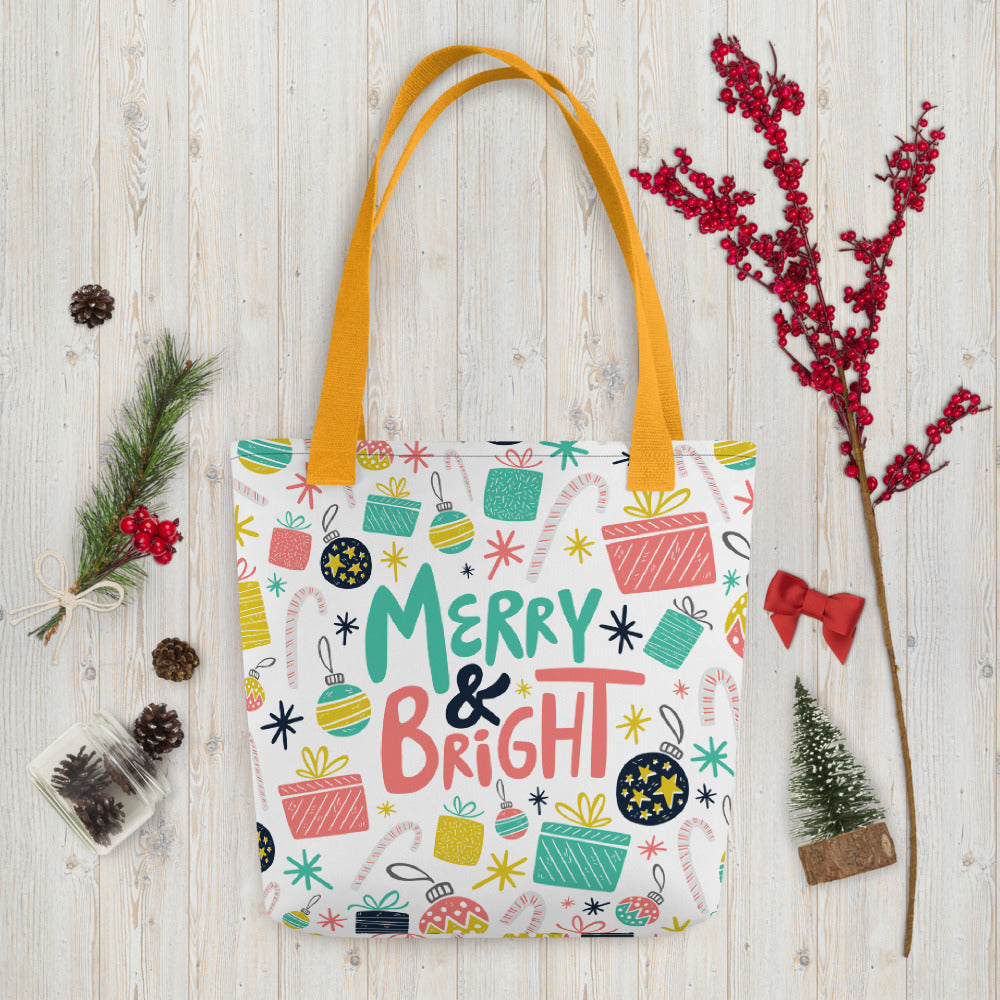 A white tote bag with yellow handles laying on a table with Christmas items around it. The tote bag features illustrated presents, ornaments, stars and candy canes. The words Merry & Bright are in the center of the pattern. The colors of the illustrated patterns are pink, light blue, yellow and black. 