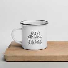 Load image into Gallery viewer, A white enamel mug sitting on top of a wood cutting board on a white background. The mug is white with the very top silver enamel. The design features hand illustrated words reading &quot;Merry Christmas&quot; with illustrated pine trees under the words. The design on the mug is in black. 