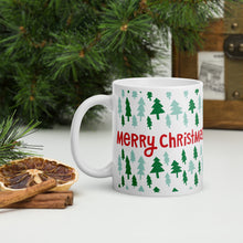 Load image into Gallery viewer, A white mug with a pine tree in the background. The mug feaures the words &quot;Merry Christmas&quot; with light and dark green pine trees around the words. The words are in red.