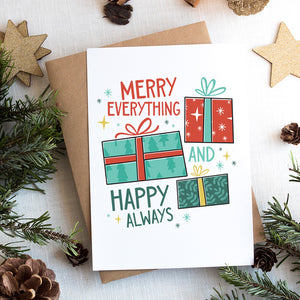 A photo of a Christmas card on top of a brown paper wrapped gift with Christmas decor around it. The card has a white background with the words "Merry Everything and Happy Always." There are three illustrated Christmas gifts in light red, green and blue with patterns on them.