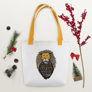 A white tote bag with mustard yellow handles laying on a white tabletop with pinecones and a red berry branch around it. The tote bag has mustard yellow handles and an illustration of the Aslan character. Inside the illustration there is the quote “At The Sound of Your Roar, Sorrows Will Be No More.” 
