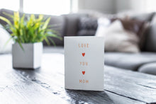 Load image into Gallery viewer, A greeting card featured on a black, wood coffee table. There’s a white planter in the background with a green plant. There’s also a gray sofa in the background with a white pillow. The card features the words “Love You Mom.” 