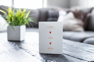 A greeting card featured on a black, wood coffee table. There’s a white planter in the background with a green plant. There’s also a gray sofa in the background with a white pillow. The card features the words “Love You Mom.” 